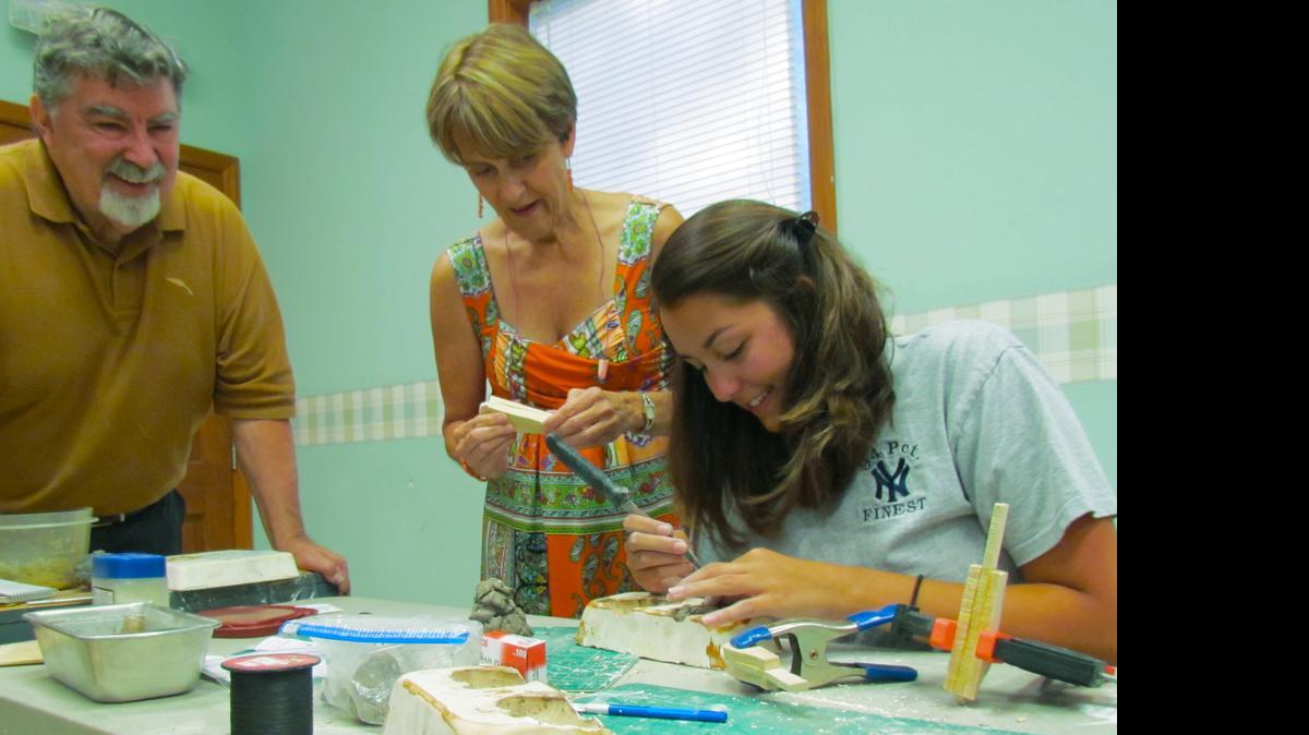 
Marionette Workshop teacher Frank Blackmon and student Debbie Skillman, who is applying pressure to seal the pieces of wood she has glued together, watch student Luci Tramposch chip away the Plaster of Paris that encases her clay mold. 
