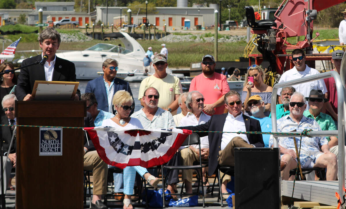 Captain Archie 'Poo' McLauchlin, wearing a blue Hawaiian shirt and dark sunglasses (right) and flanked by State Senator Greg Hembree to his  left, listens as State Representative Greg Duckworth speaks during the dedication to rename the Little River Swing Bridge after Captain Poo.