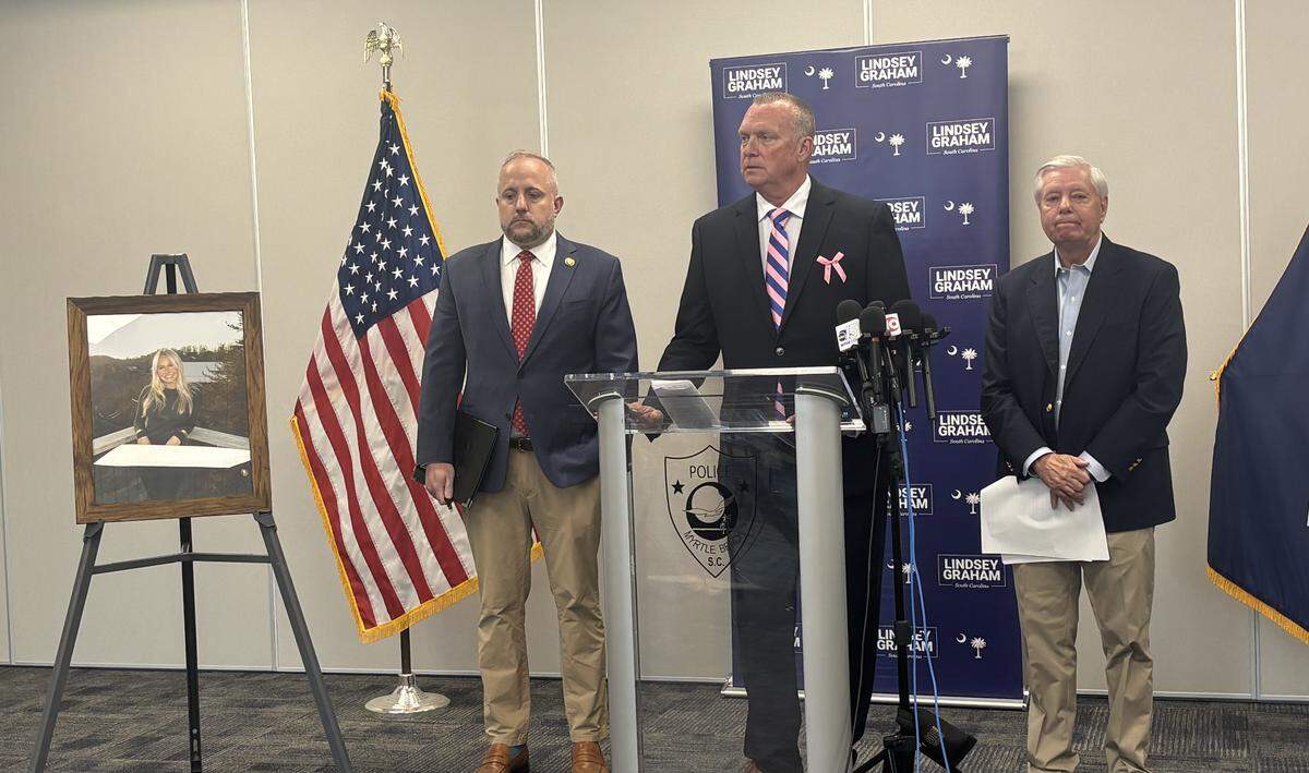 Three men wearing suits stand in front of a stand with microphones attached to it. Behind them is a blue banner. To the left is a picture of a blonde woman smiling.