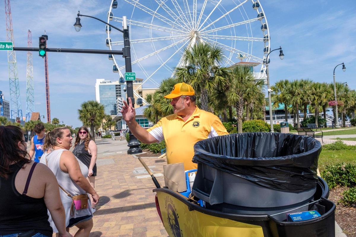 Jeremy Trendle waves to tourists walking along Ocean Boulevard in Myrtle Beach on Wednesday afternoon answering questions. A dozen Gold Cap Ambassadors are now patrolling the streets of Myrtle Beach cleaning up trash, providing hospitality services, and reporting issues to city officials.The pilot program started in August as a way to improve the safety and cleanliness of the tourism district. August 26, 2020.