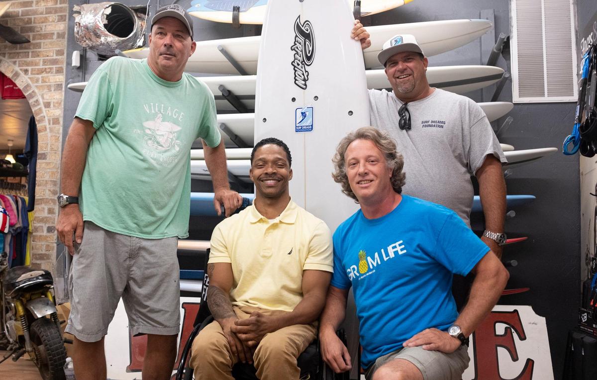 Kelly Richards (left), along with Tyler Watkins (bottom right) and Phil Jackson (top right) presnt quadriplegic surfer Ernie Johnson with a new board given by the Surf Dreams Foundation and shaped by Richards. Johnson will be competing in the USA Surfing adaptive Championships on June 13, 2019.