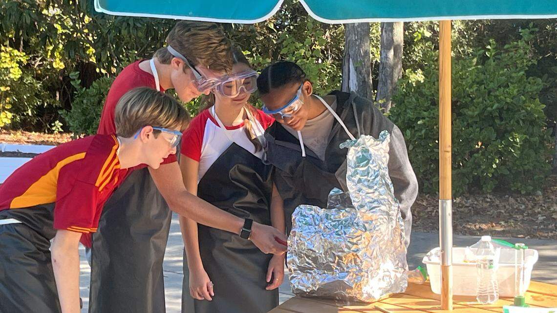 Four students all wearing protective gear stand underneath an umbrella while working on a class project.