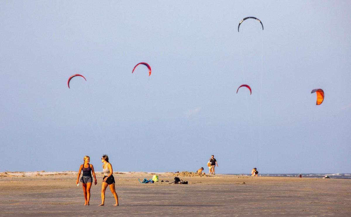 Beachgoer walk the broad expanse of the Sullivan’s Island beach front as kiteboarder’s sails fly in the distance. Shifting sands are having opposite effects on some Charleston area beaches with erosion claiming the Folly Beach shoreline while Sullivan Island’s beach is growing. July 21, 2022.
