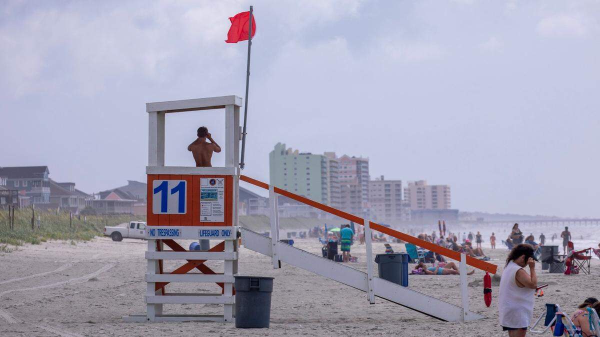 A North Myrtle Beach lifeguard looks looks through binoculars at swimmers along Cherry Grove Beach as a red flag waves warning of dangerous conditions. June 23, 2023.