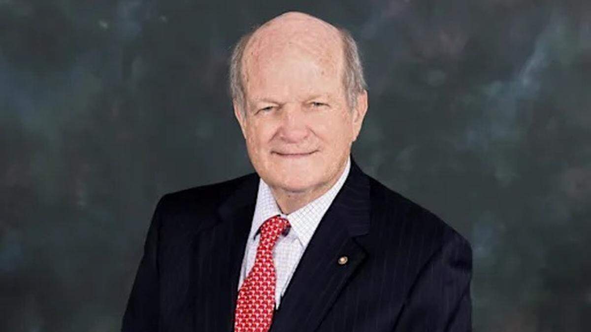 A man poses for a photo in front of a gray backdrop. He is wearing a suit with a red tie.