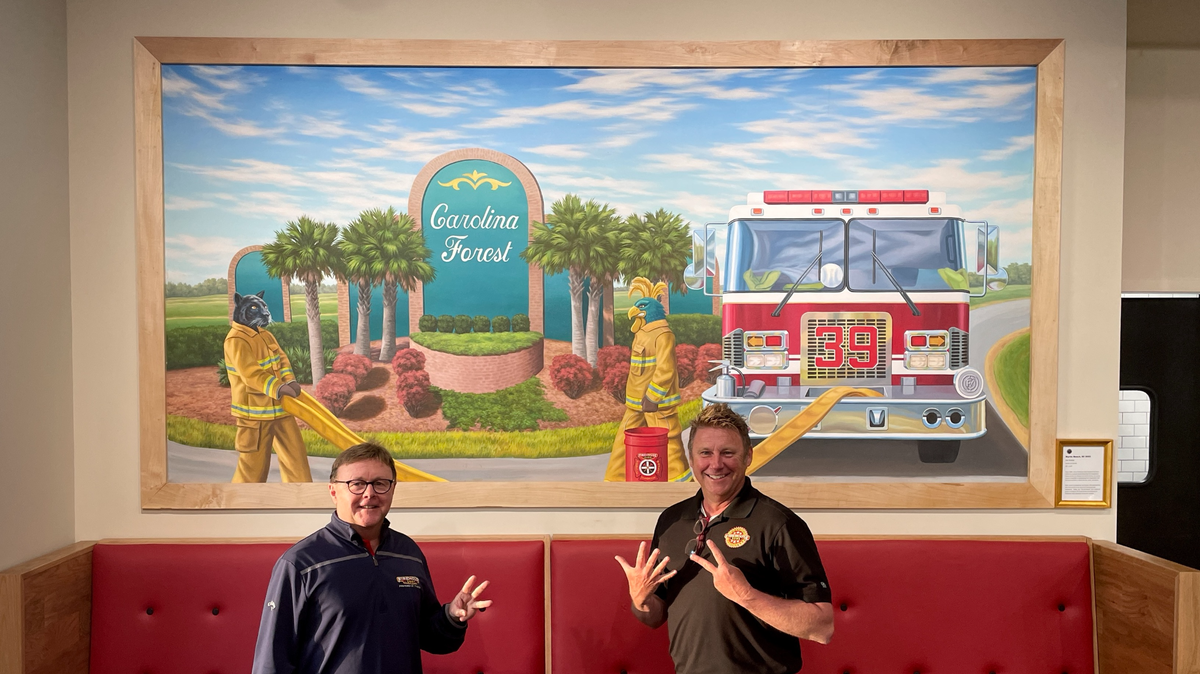Larry Chandler and Jody Chandler stand in front of their new Myrtle Beach restaurant’s hand-painted mural depicting popular sports team mascots Sir Purr and Chauncey dressed as firefighters next to a local fire truck with the Carolina Forest welcome sign in the background.