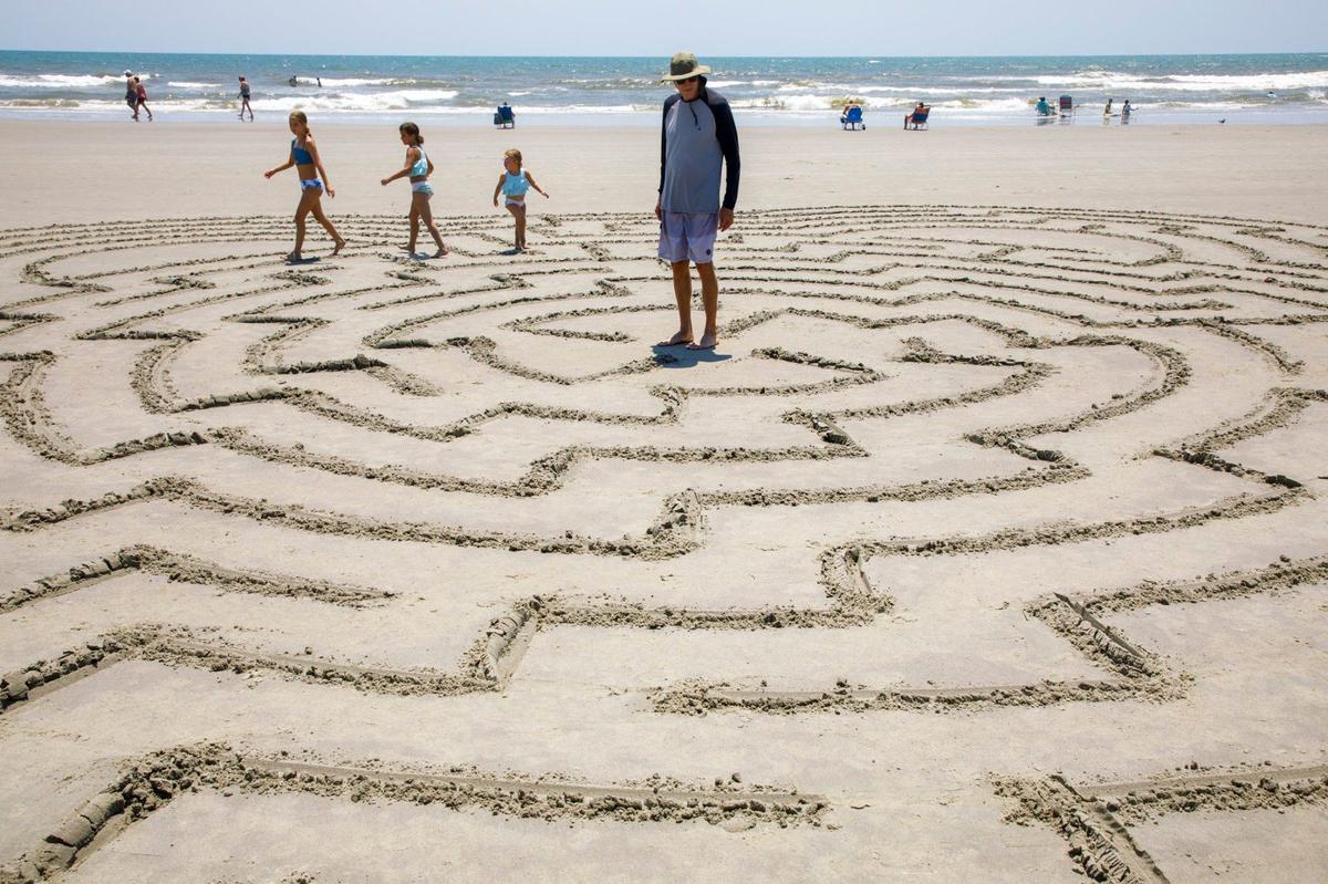 Dave Pavlick, of Canton, OH stands in the middle of a sand maze on Sunset Beach, N.C. Hunter Gibbes, known locally as “Sunset Beach Maze Man,” has been making mazes at Sunset Beach, N.C. for nearly 20 years. The sand artist makes up the designs on the spot. Vacationers and locals rush to complete the mazes he creates before the tide comes in and creates a clean slate for his next artwork. Aug. 1, 2024.
